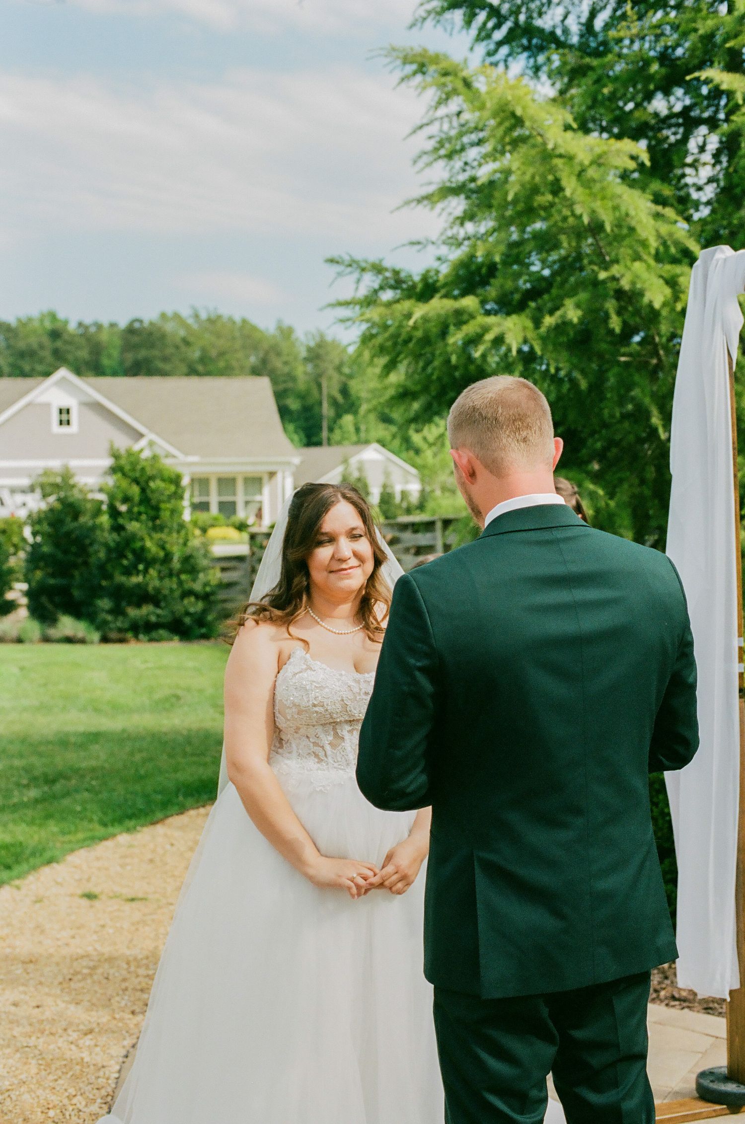 Bride smiling at groom during outdoor wedding ceremony, wearing lace gown and veil, with green trees and house backdrop.