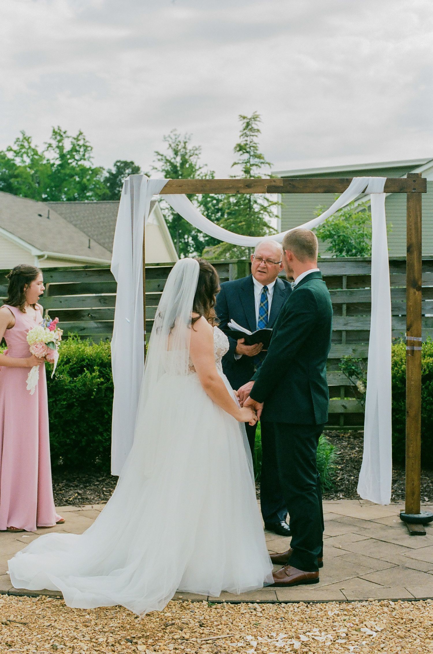 Outdoor wedding ceremony with bride in white gown and groom in navy suit exchanging vows under a draped wooden arch.