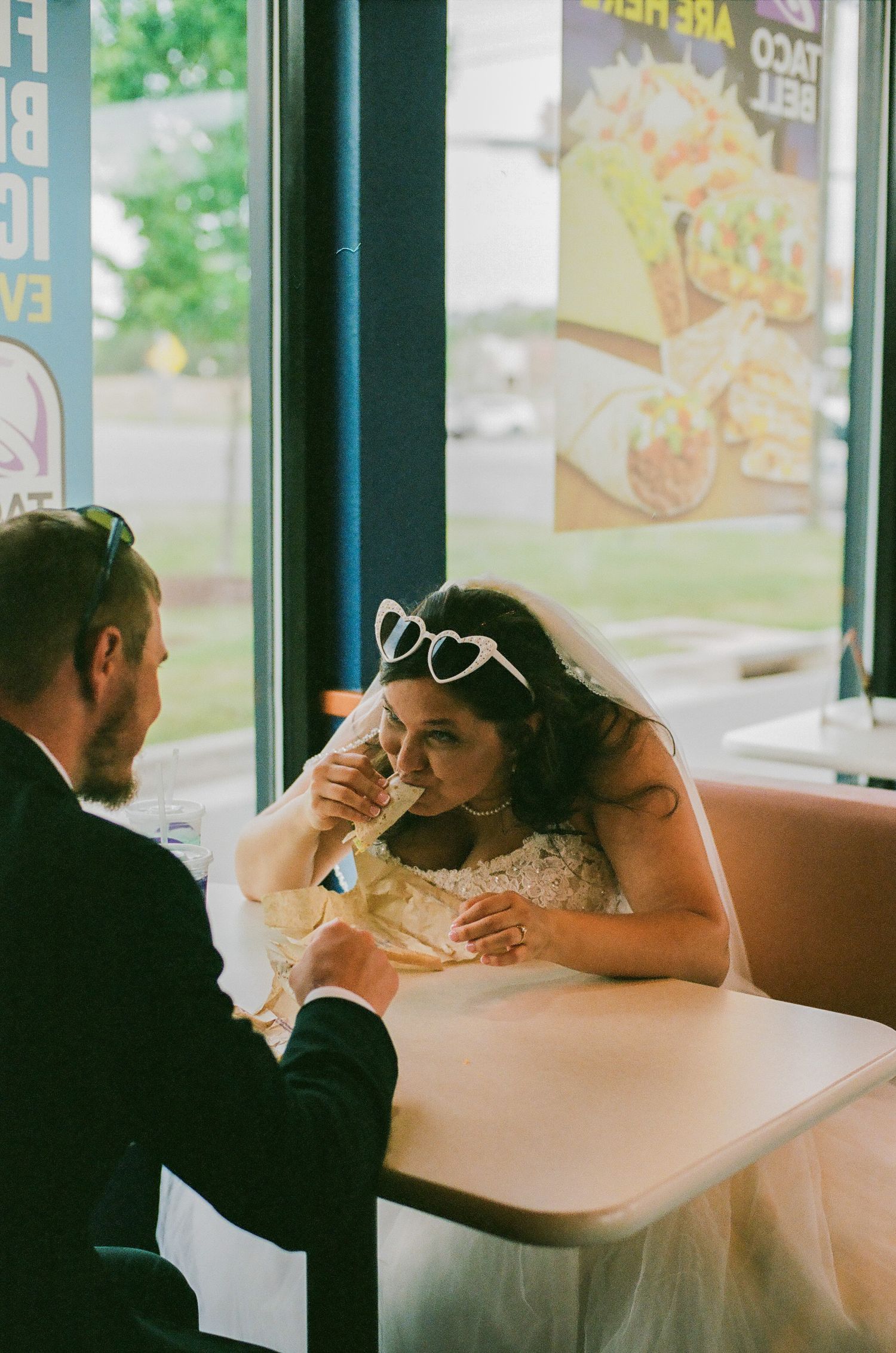 Bride in wedding dress and heart sunglasses drinking at Taco Bell with groom in black suit at booth.