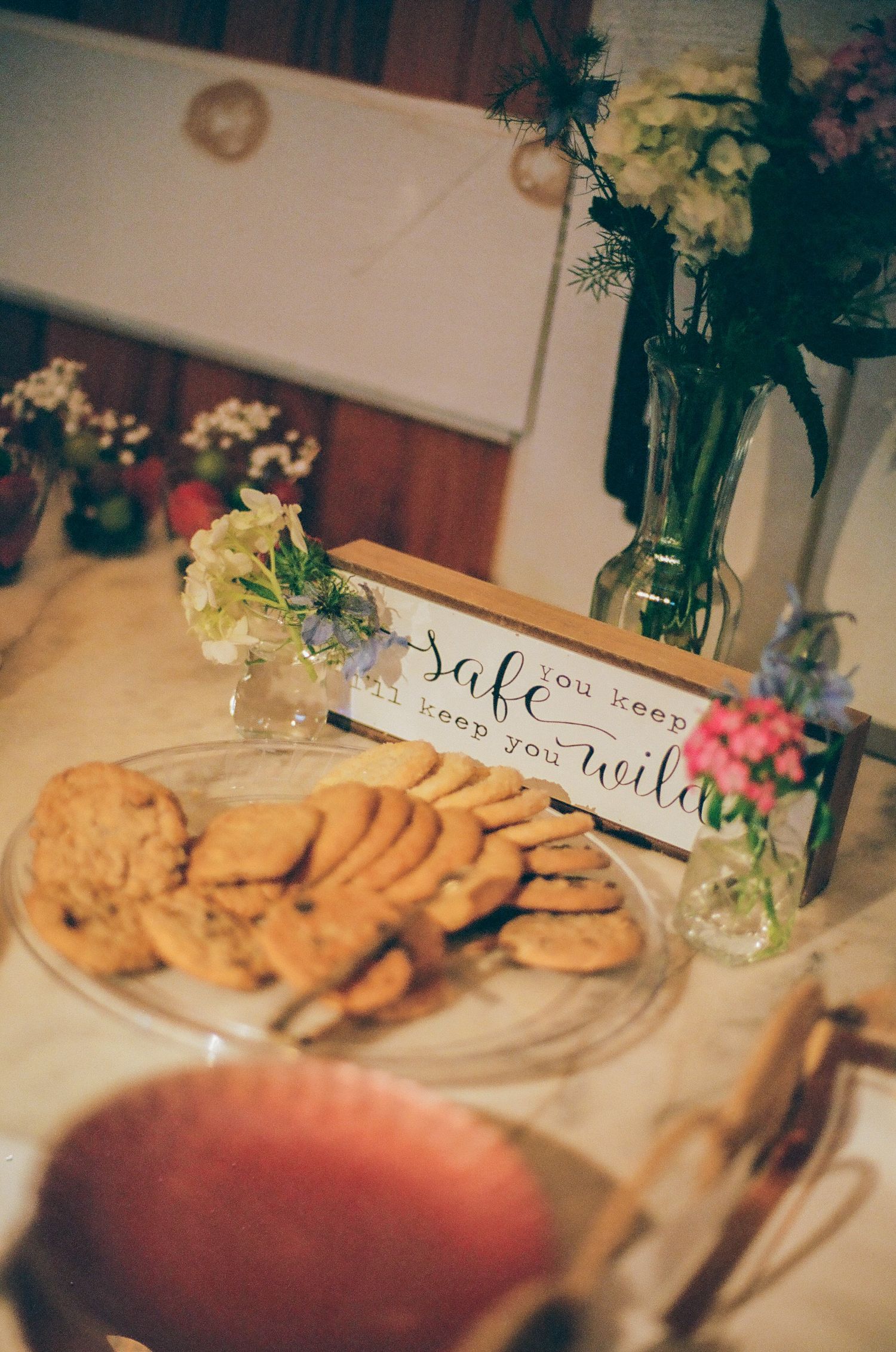 Rustic dessert table with cookies on glass plate, floral arrangements, and a decorative sign at a wedding reception.