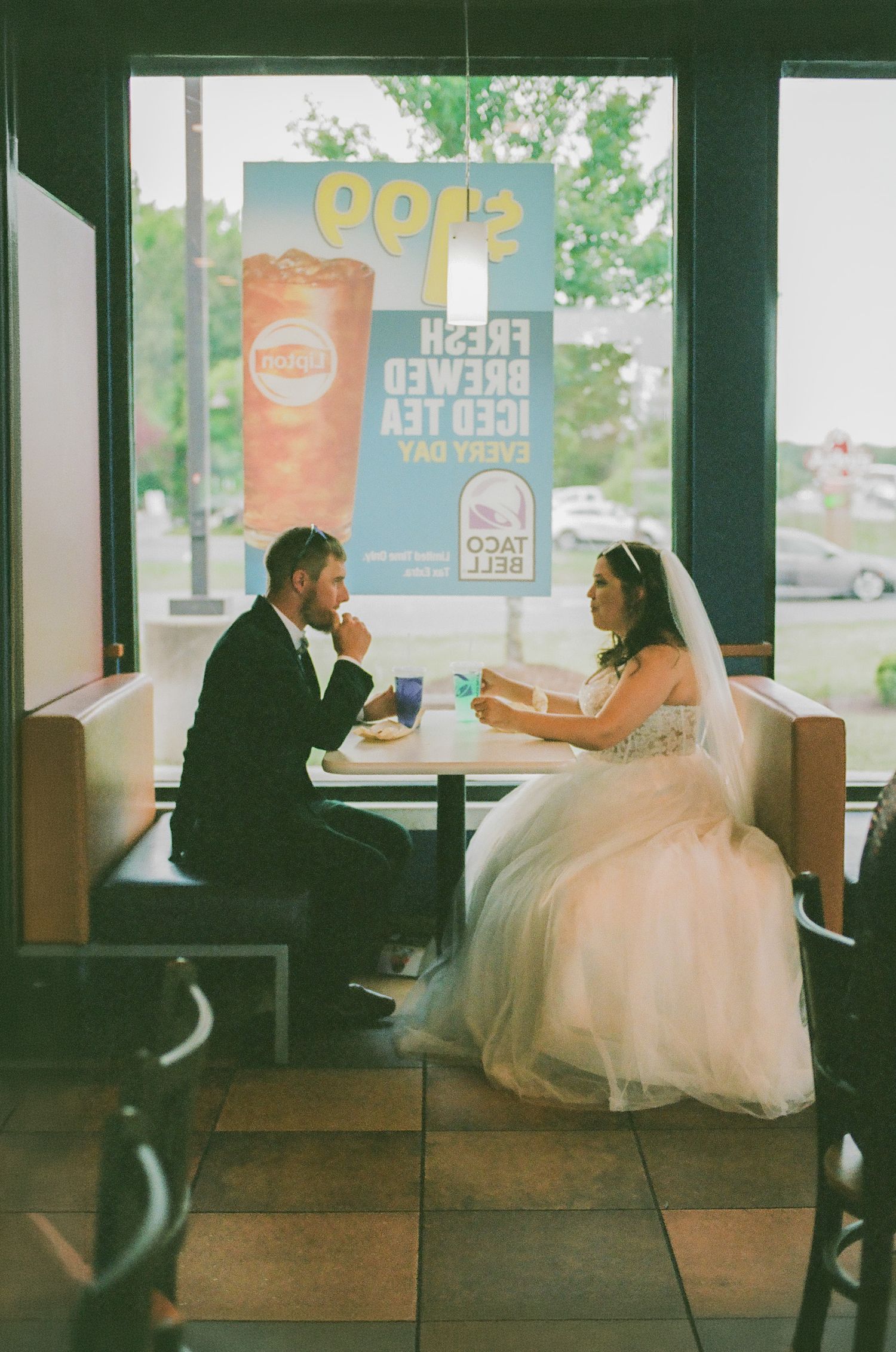 Bride and groom in wedding attire sharing a romantic meal at Taco Bell, sitting in a booth with a Fresh Brewed Iced Tea sign.