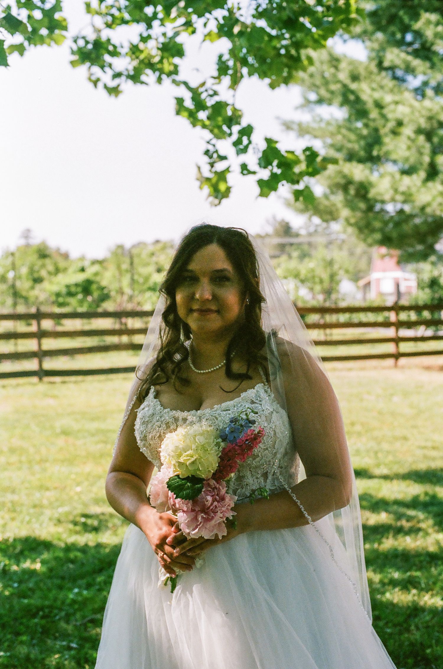 Bride in white lace gown and veil holds colorful bouquet outdoors near wooden fence on sunny wedding day.