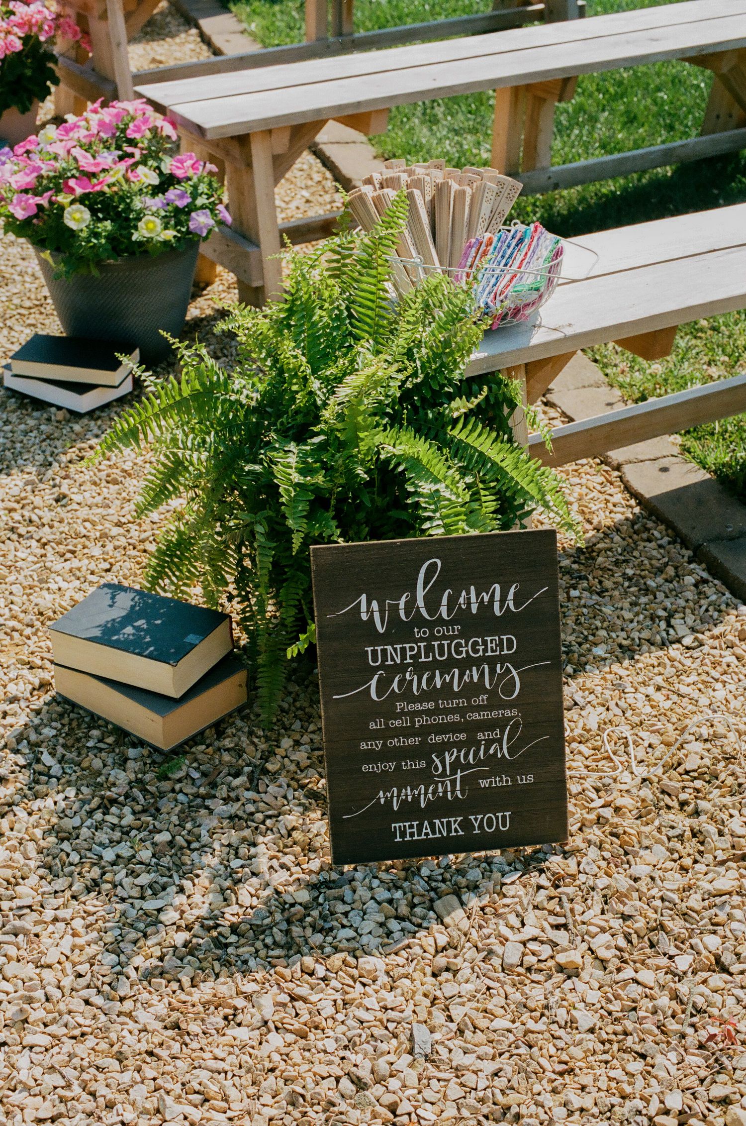 Rustic wedding unplugged ceremony sign surrounded by ferns and flowers on gravel with wooden benches in background.