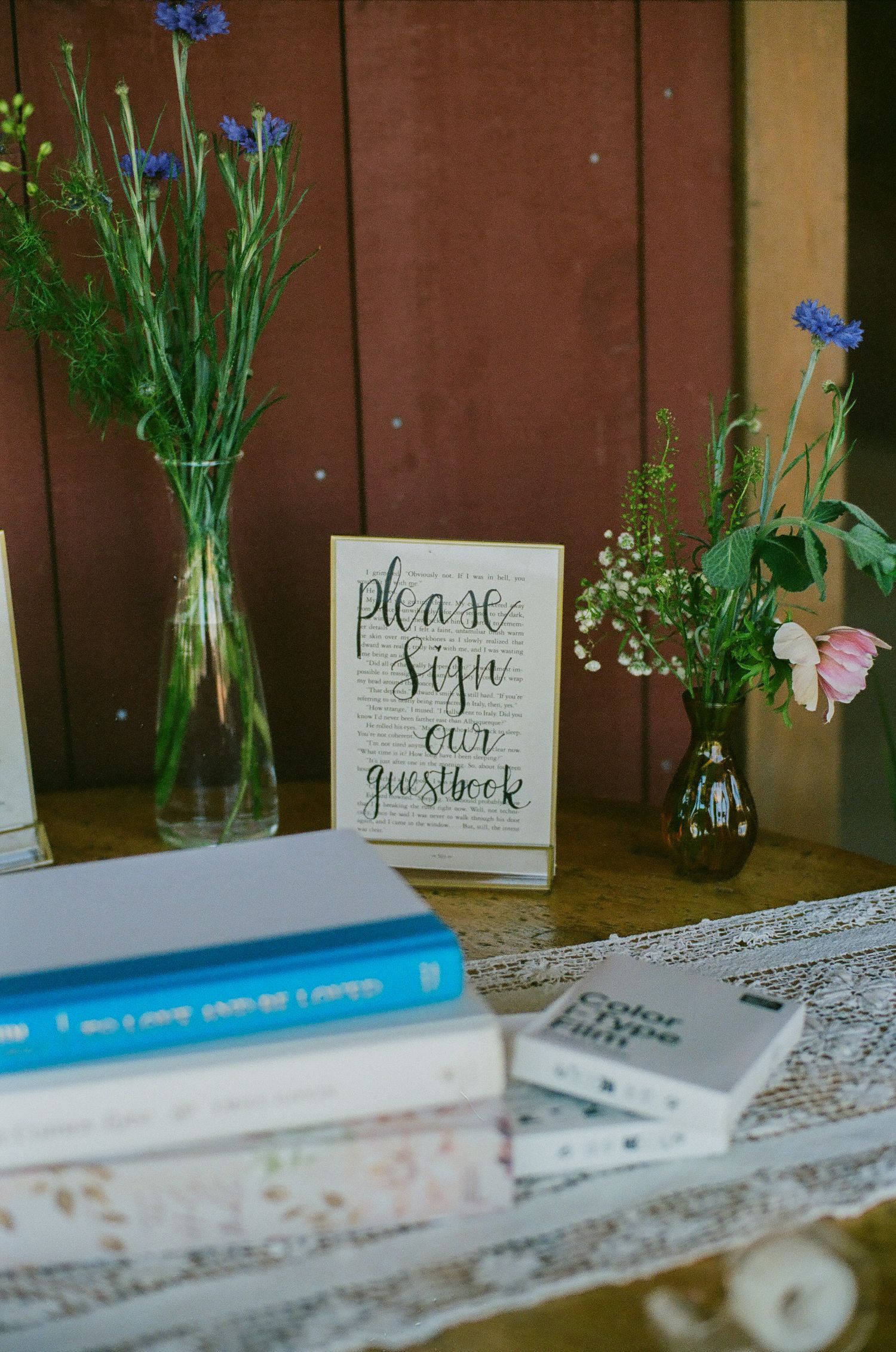 Wedding guestbook sign displayed with wildflowers in glass vases and stacked books on a rustic lace-covered table.