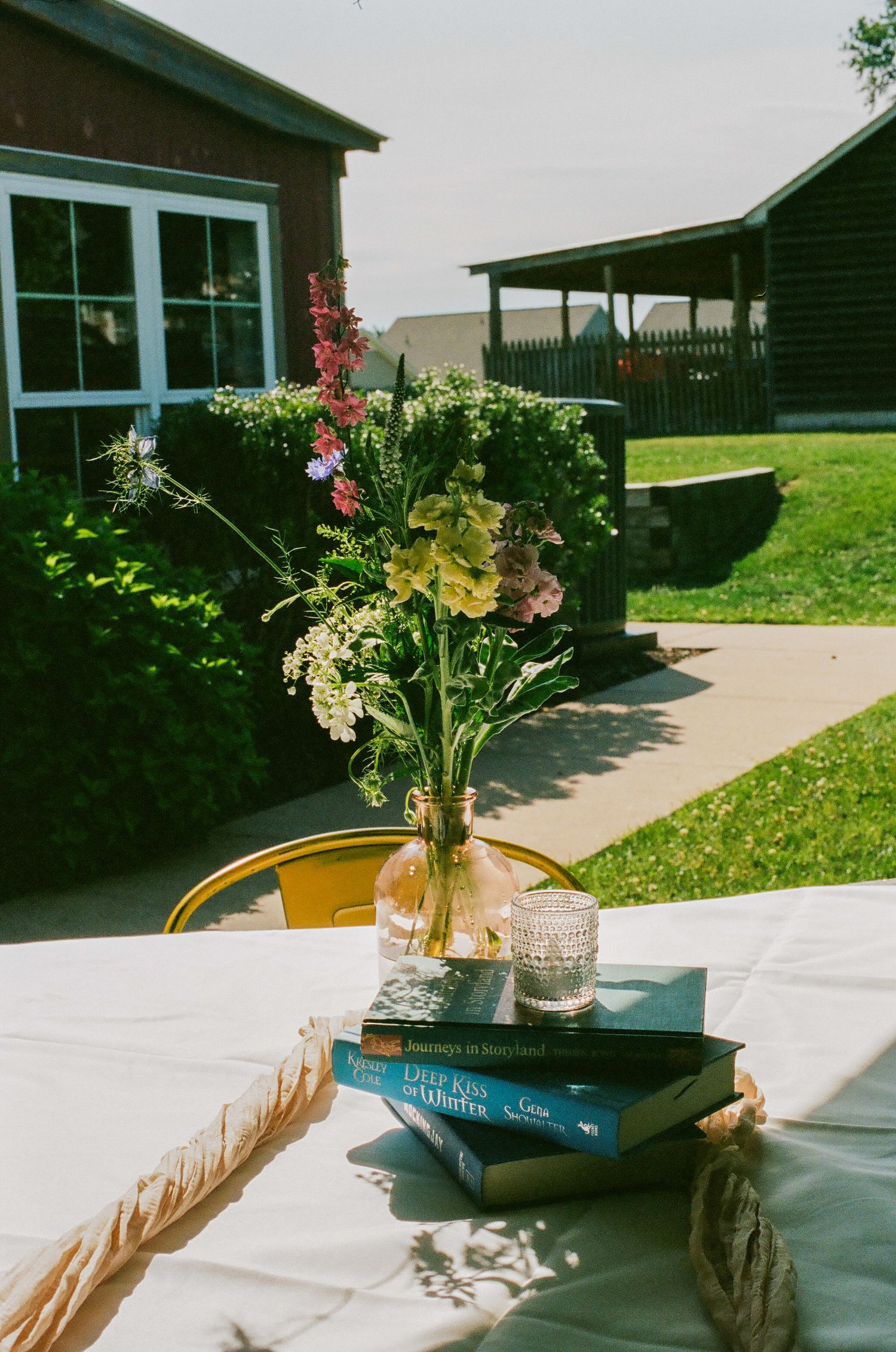 Rustic outdoor table with wildflower bouquet in glass vase, stacked teal books, and rope detail, set against a barn backdrop.