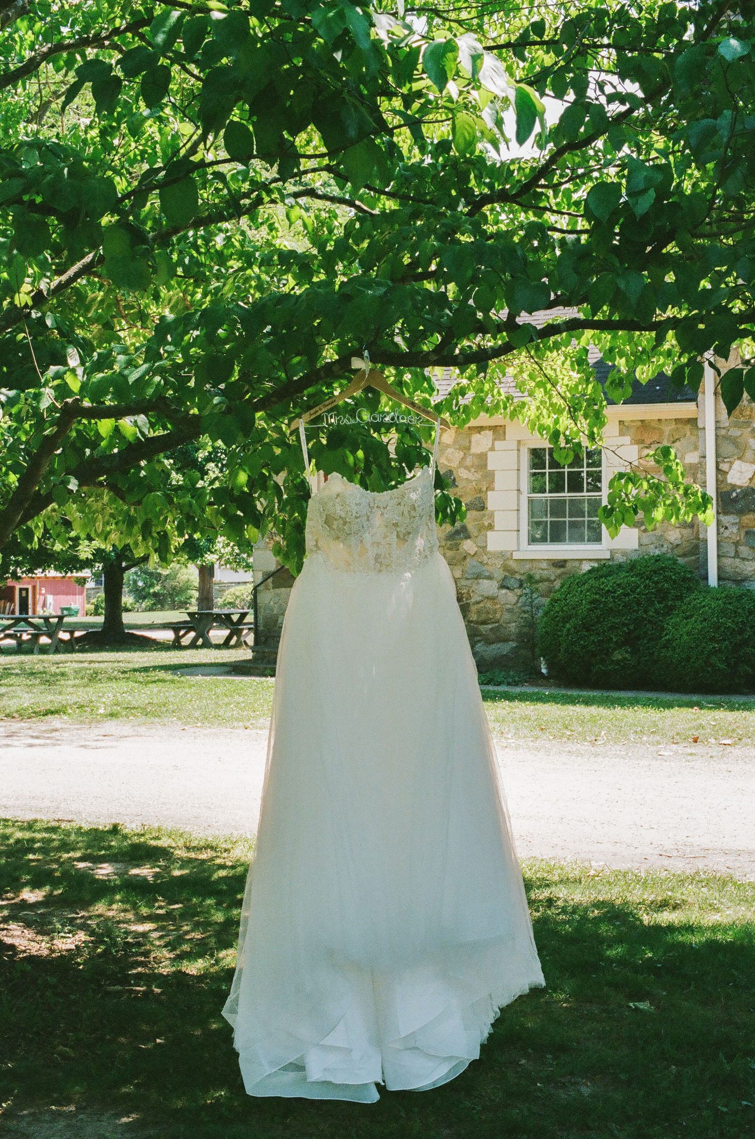 A white lace wedding dress hanging from a tree branch outdoors with a stone building in the background.