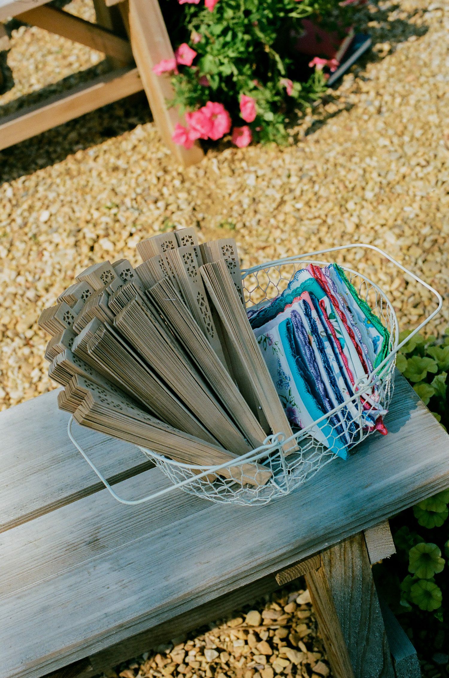 White wire basket holding wooden hand fans and colorful fabric fans on a wooden bench outdoors.