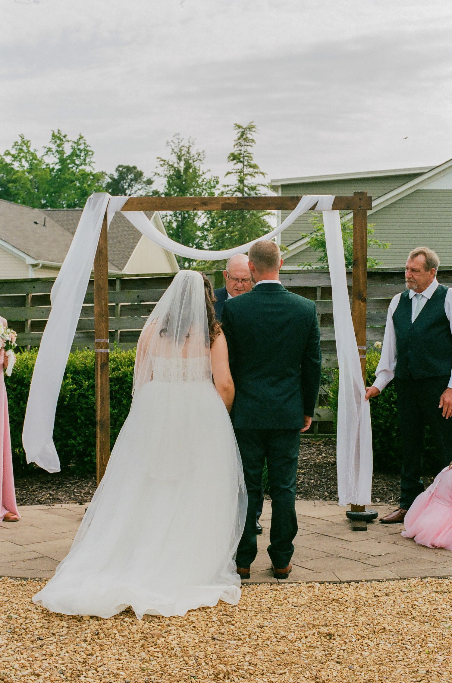 Bride and groom stand before wooden arch with white draping during outdoor wedding ceremony.