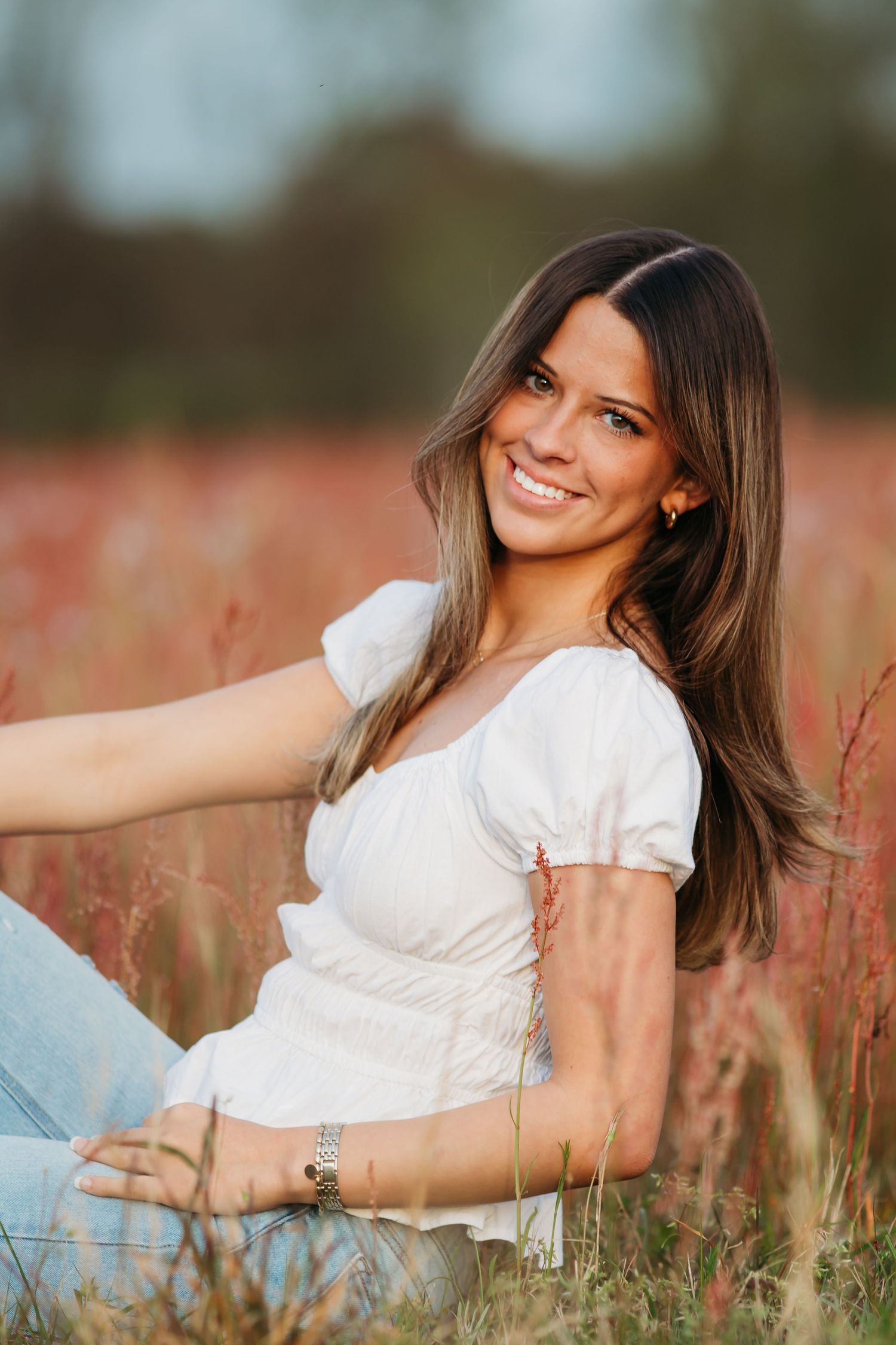 A smiling woman in a white top sits in a field of tall reddish grass during sunset.