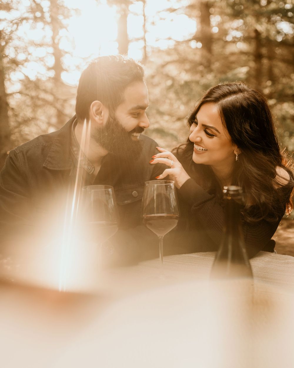A romantic couple shares wine and laughter during a sunset photoshoot in a forest setting.