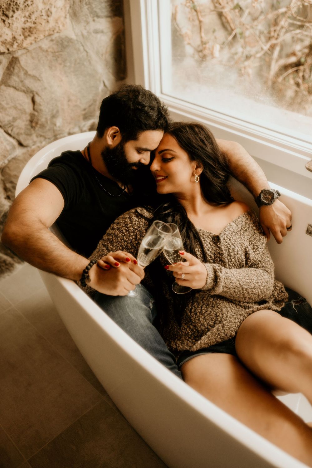 Couple sharing tender moment while drinking wine together in vintage bathtub next to window.