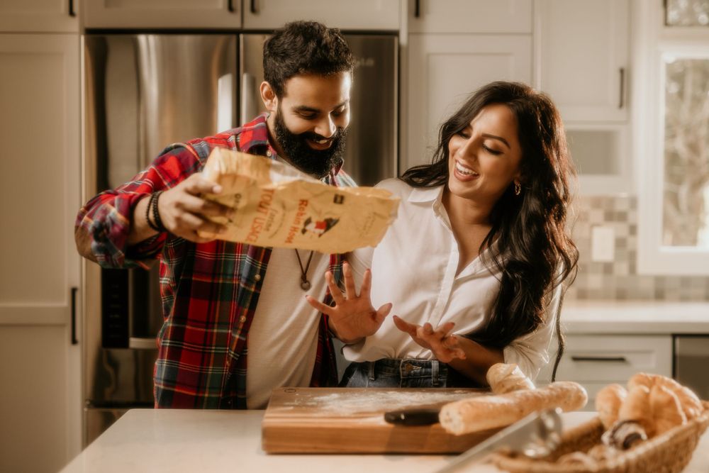 Two people laughing while sharing bread in a rustic kitchen over a wooden cutting board.