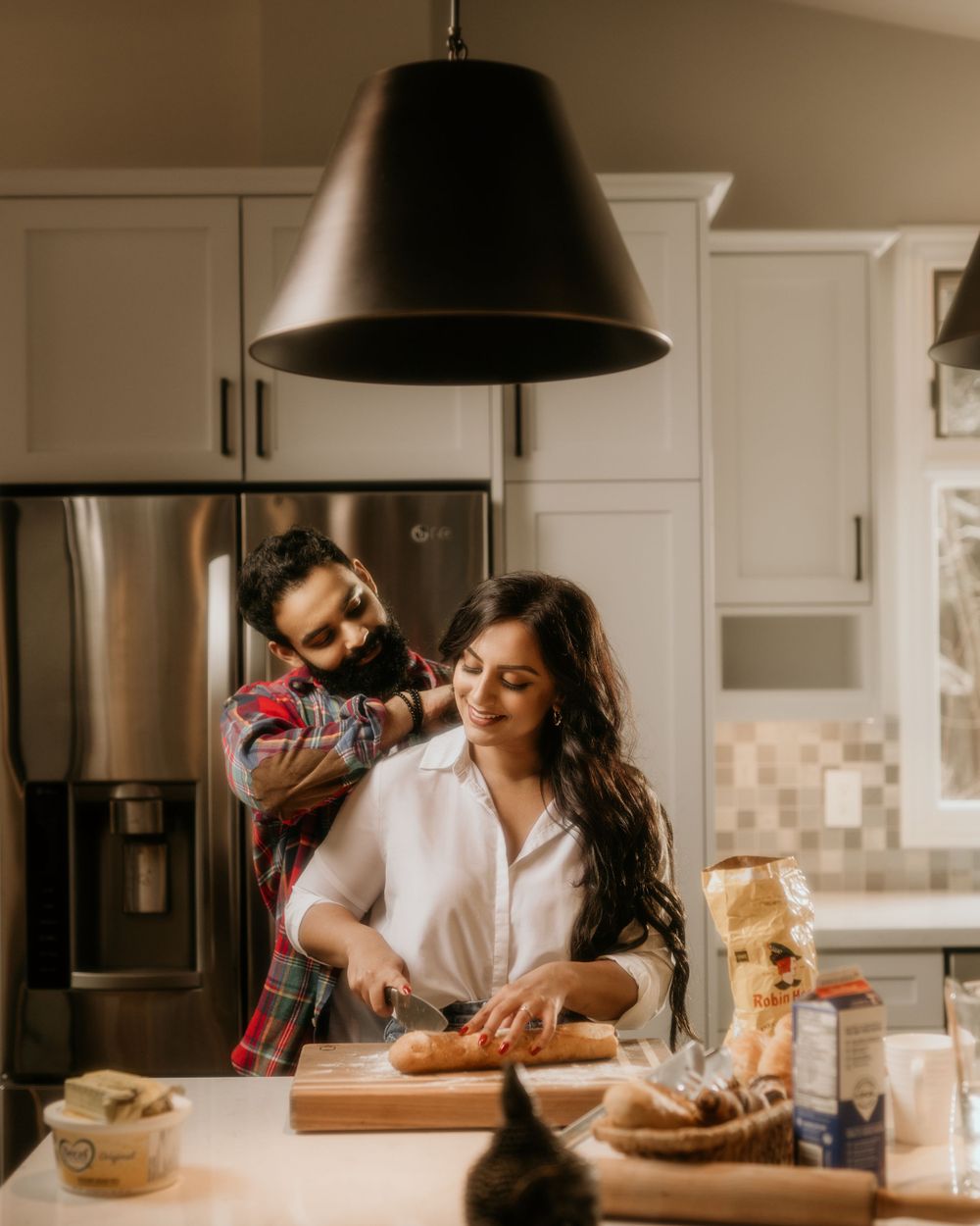 Couple cooking together in modern kitchen with black pendant lamp and stainless steel refrigerator.