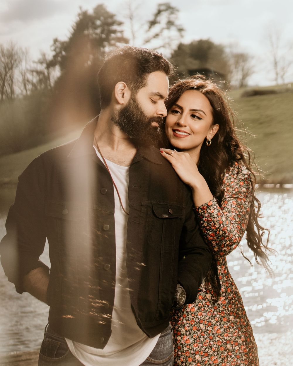 A couple shares a tender moment by the water, with soft sunlight filtering through trees in a romantic outdoor setting.