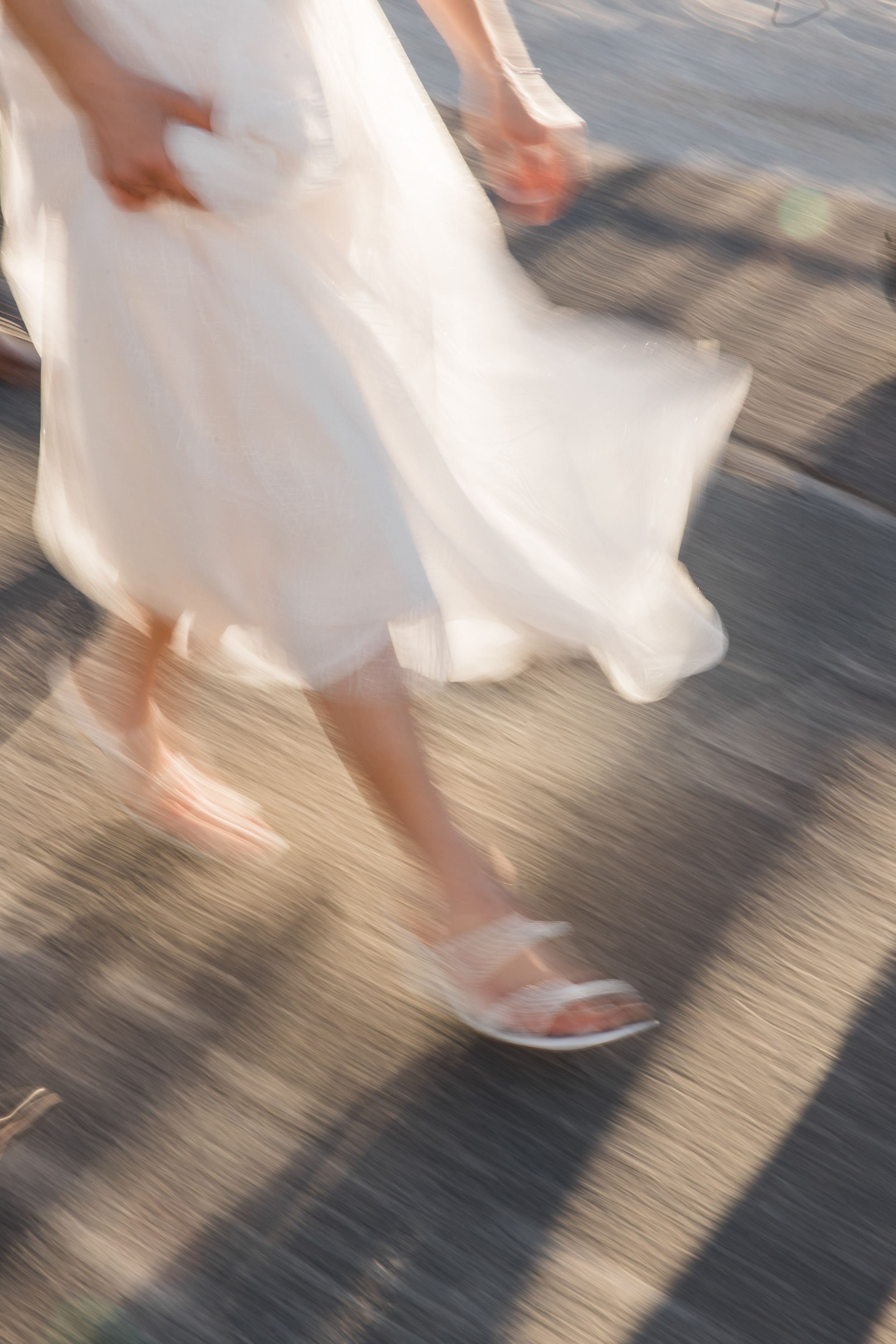 Motion blur image of white wedding dress and sandals walking on pavement.