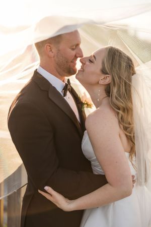 Romantic wedding portrait of newlyweds sharing an intimate moment under flowing veil at sunset.