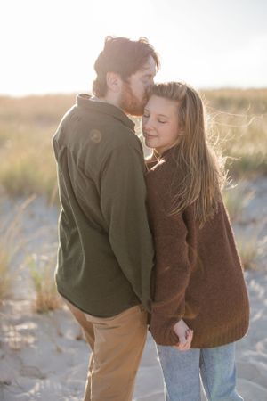 Couple sharing tender moment in warm sweaters on beach during sunset.