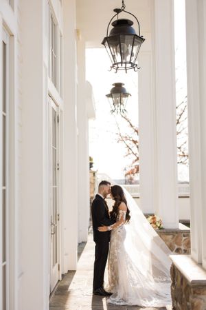 Elegant wedding couple embracing in white colonnade with vintage lanterns overhead.