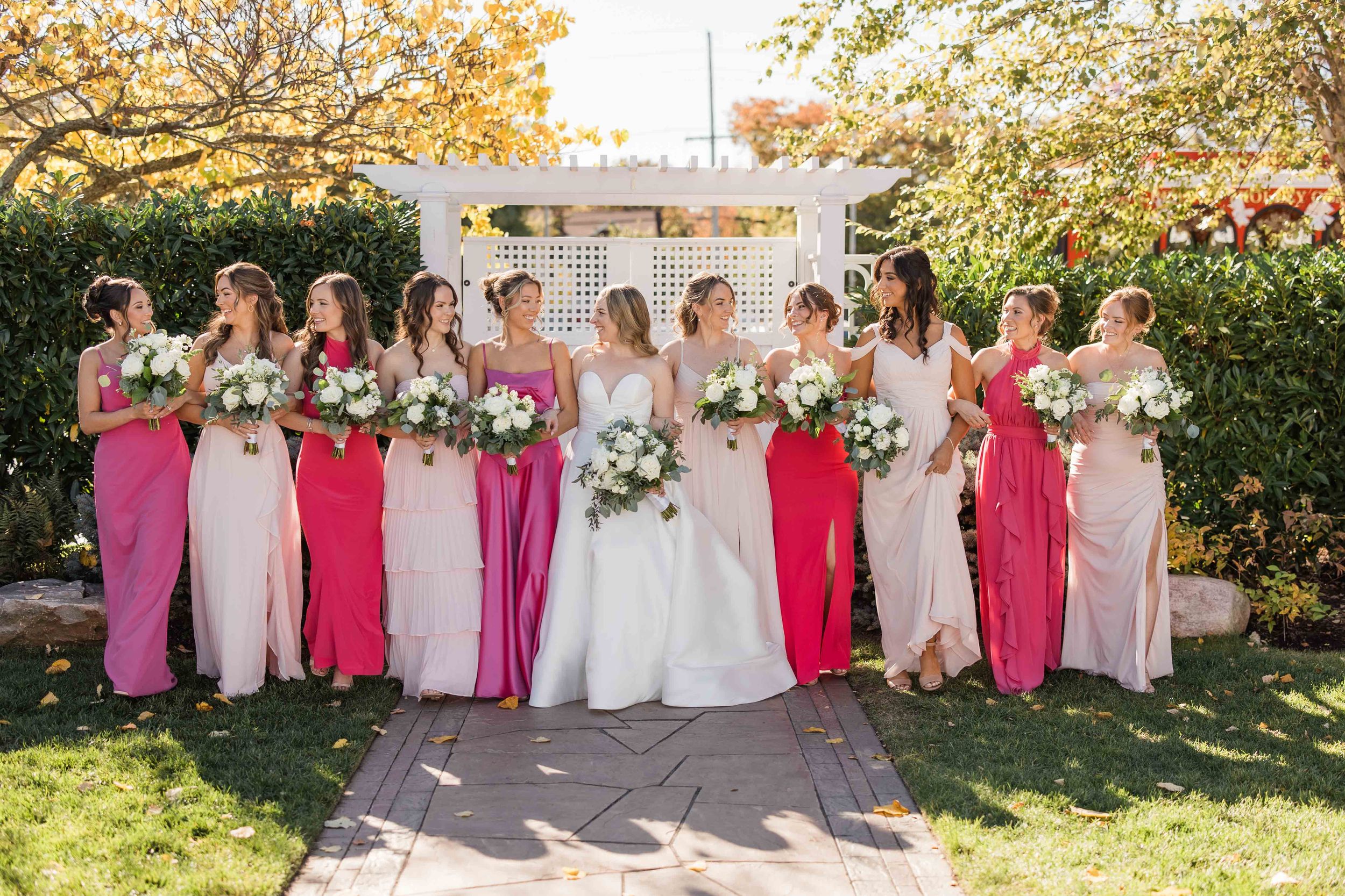 Wedding party in pink and cream colored dresses poses together in a sunny garden setting with floral bouquets.