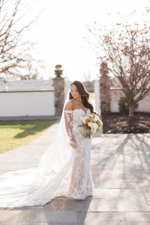 A bride in a white lace gown with long sleeves poses outdoors on a sunny day with a bouquet of flowers.