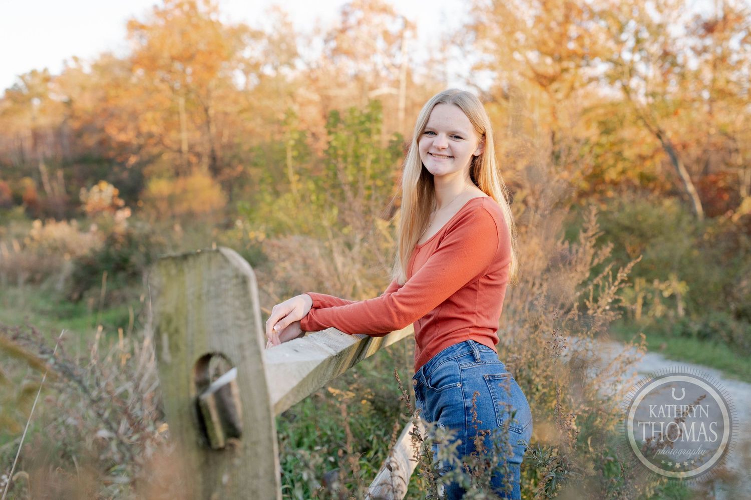 Someone in a rust-colored top leans against a wooden fence rail with autumn trees in the background.