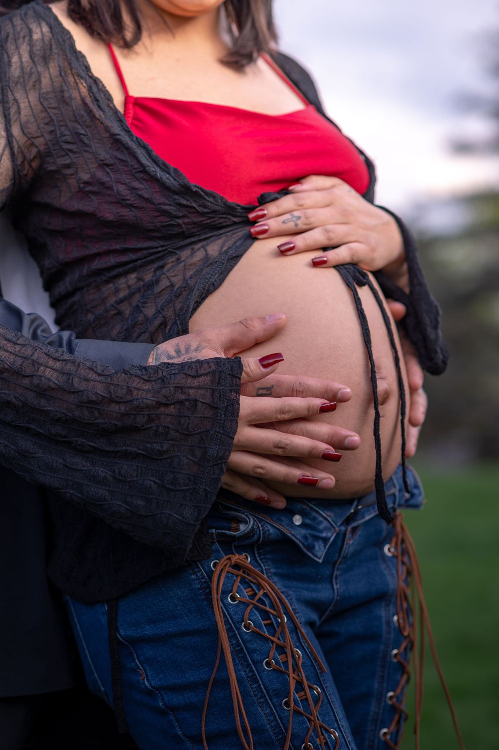 A pregnant belly is cradled by hands wearing burgundy nail polish, with a black lace top and red shirt visible.