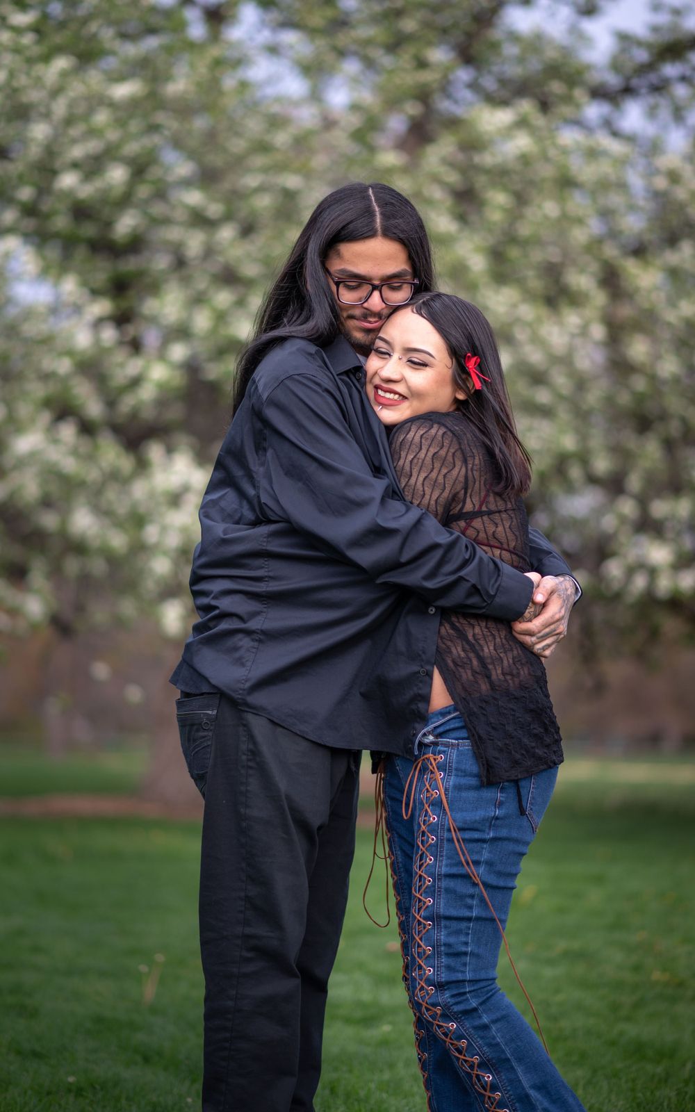 A loving embrace shared between two people in a park with blooming trees and green grass in the background.