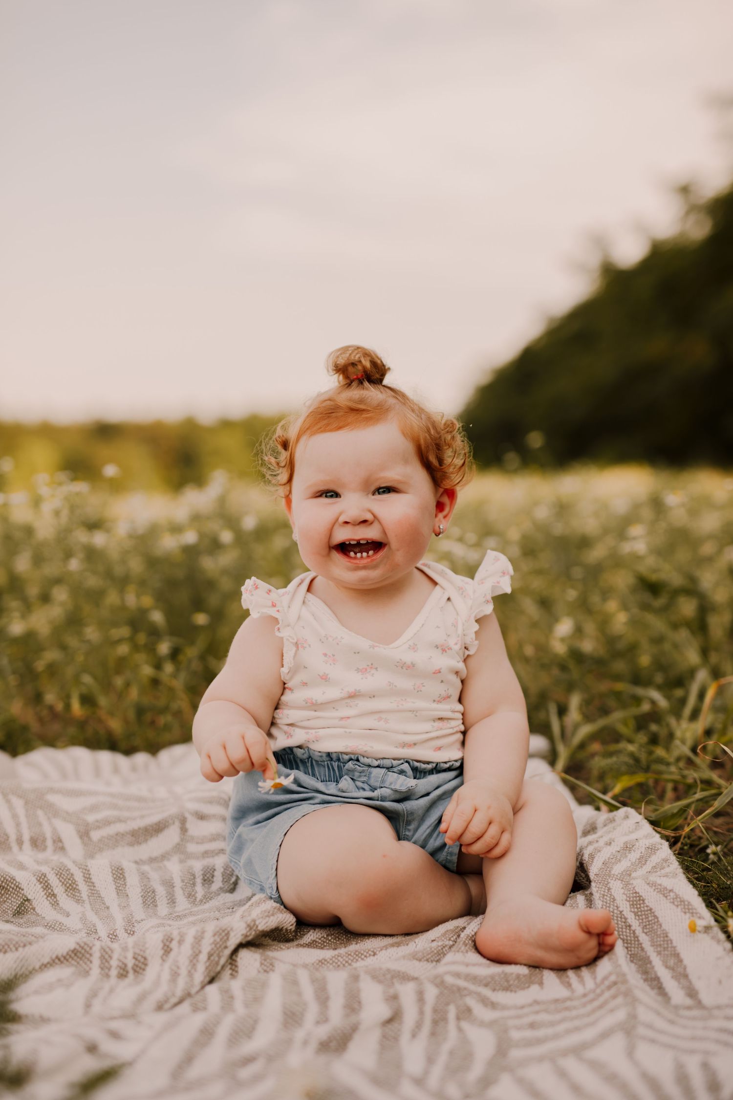 Happy baby wearing denim overalls sits on blanket in wildflower meadow during golden hour.