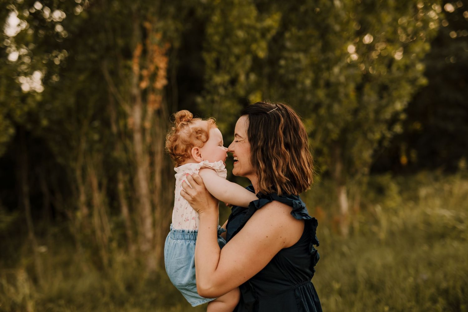 In a golden sunlit field, a person in a black tank top holds a smiling toddler wearing light blue overalls.