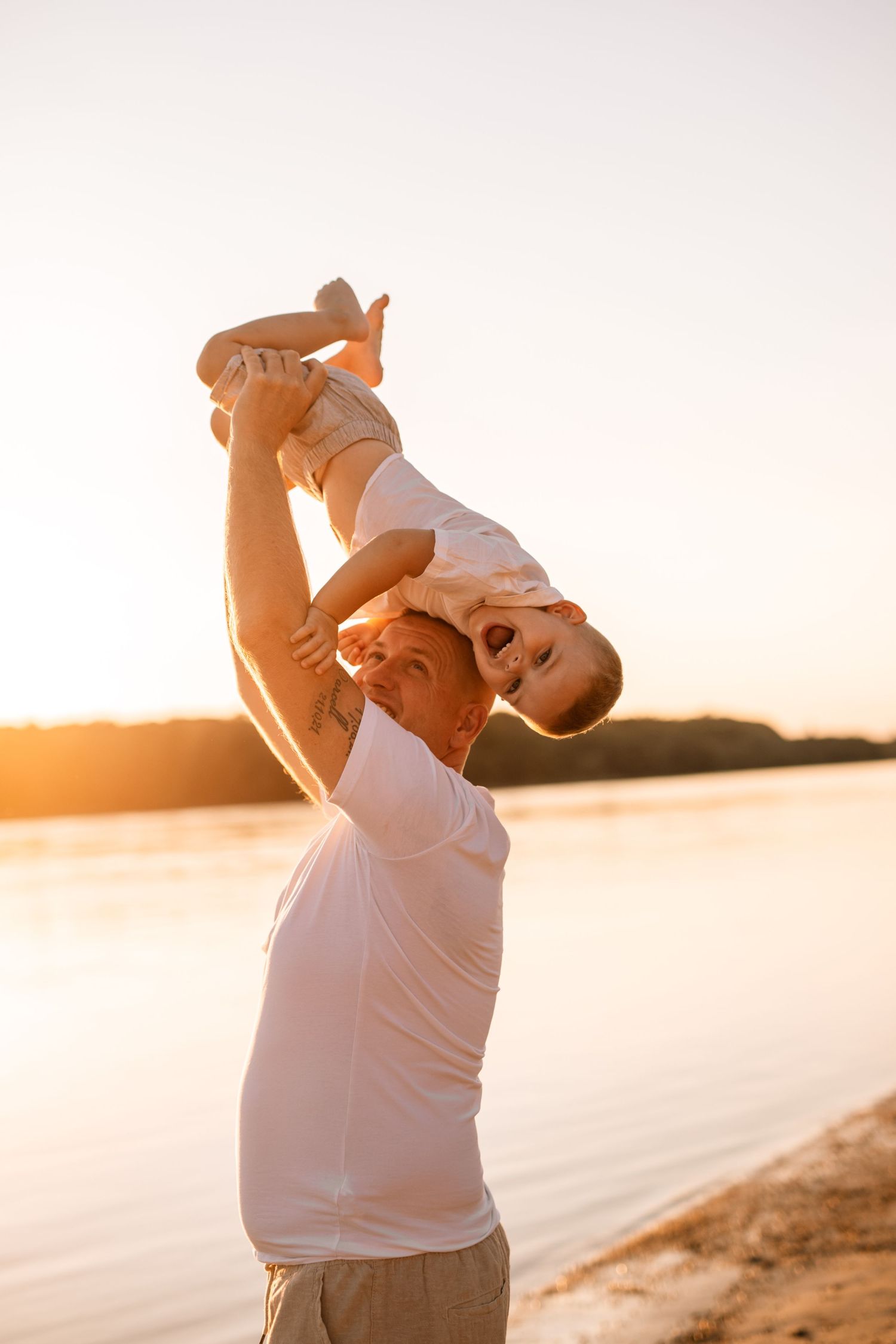 Parent holding up and playing with child along lakeside beach at sunset in warm glowing light.
