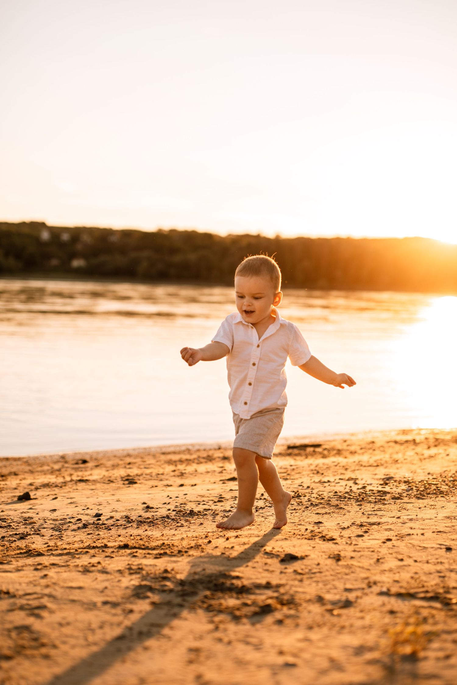 Small child playing and jumping on a beach at sunset in summer white outfit casting long shadow on sand.