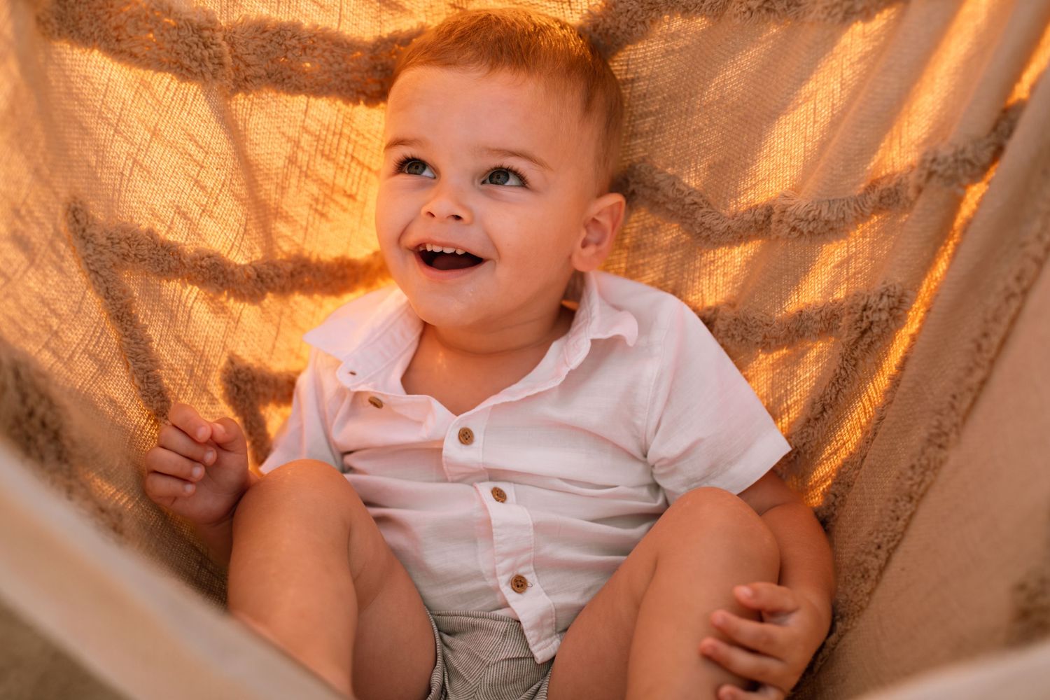 A cheerful toddler wearing a white button-up shirt sits and laughs against a gold-toned backdrop.