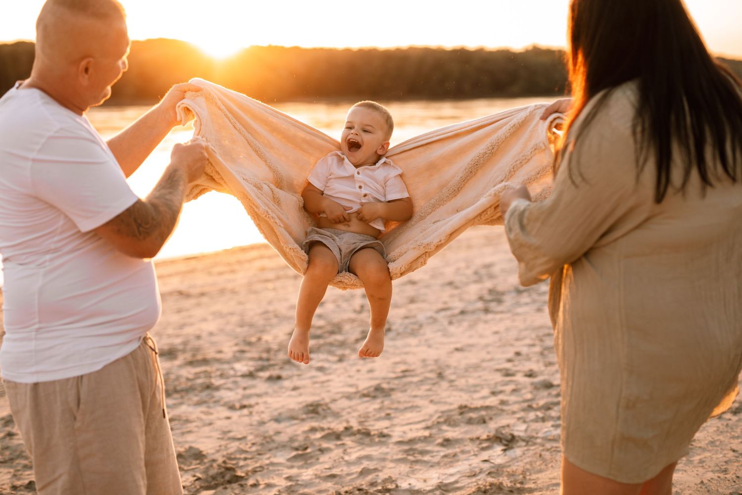 Two people hold blanket while swinging happy baby at sunset on sandy beach during golden hour.