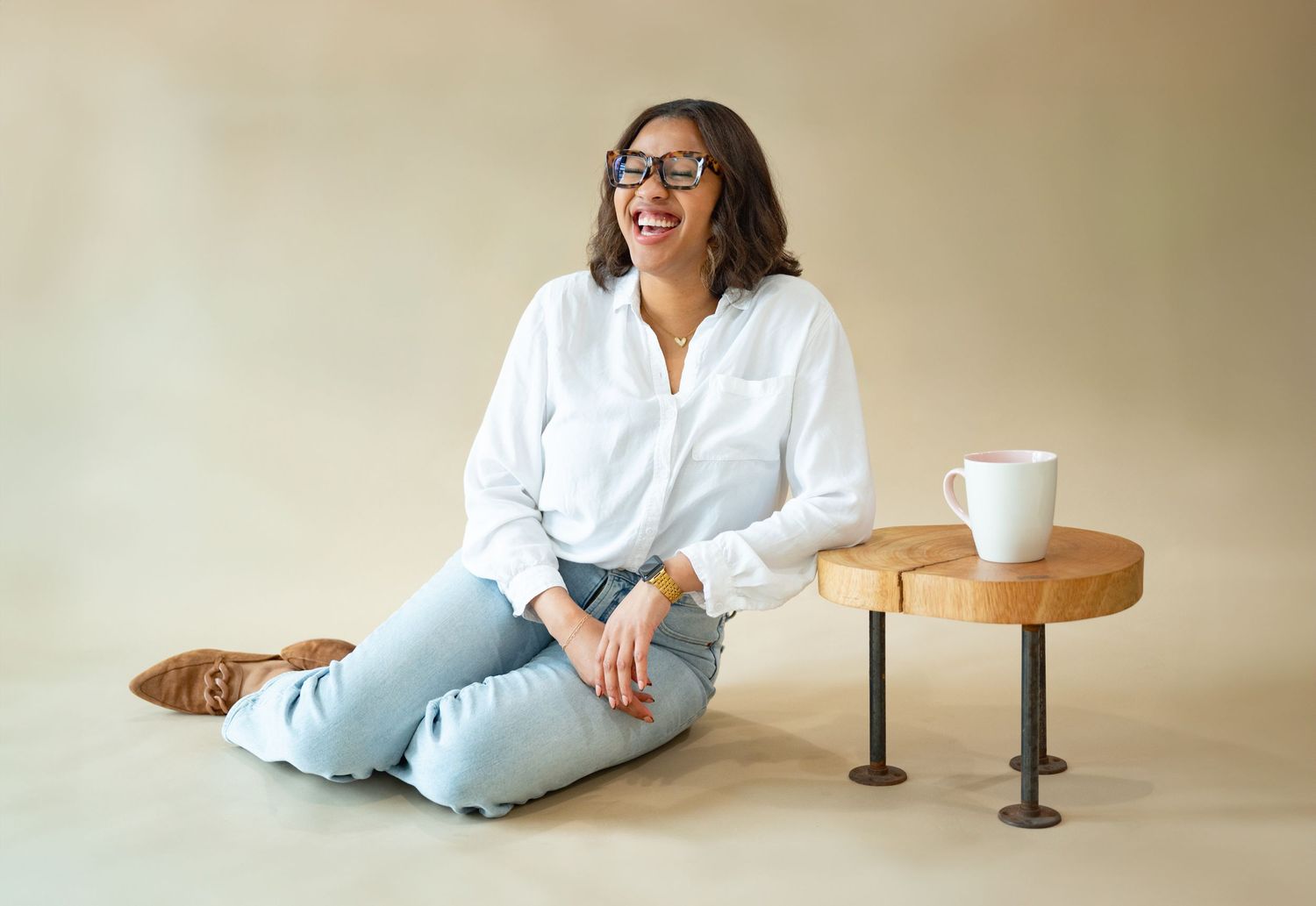 Someone wearing a white button-down shirt and jeans sits casually on the floor next to a small wooden side table with coffee mug.