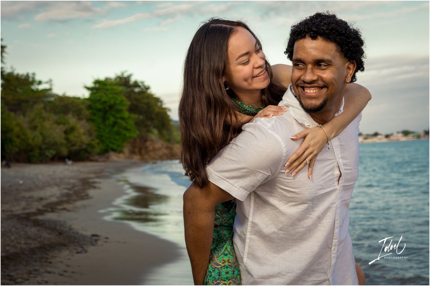 A loving couple shares a joyful embrace on a scenic beach at sunset with ocean and greenery in the background.