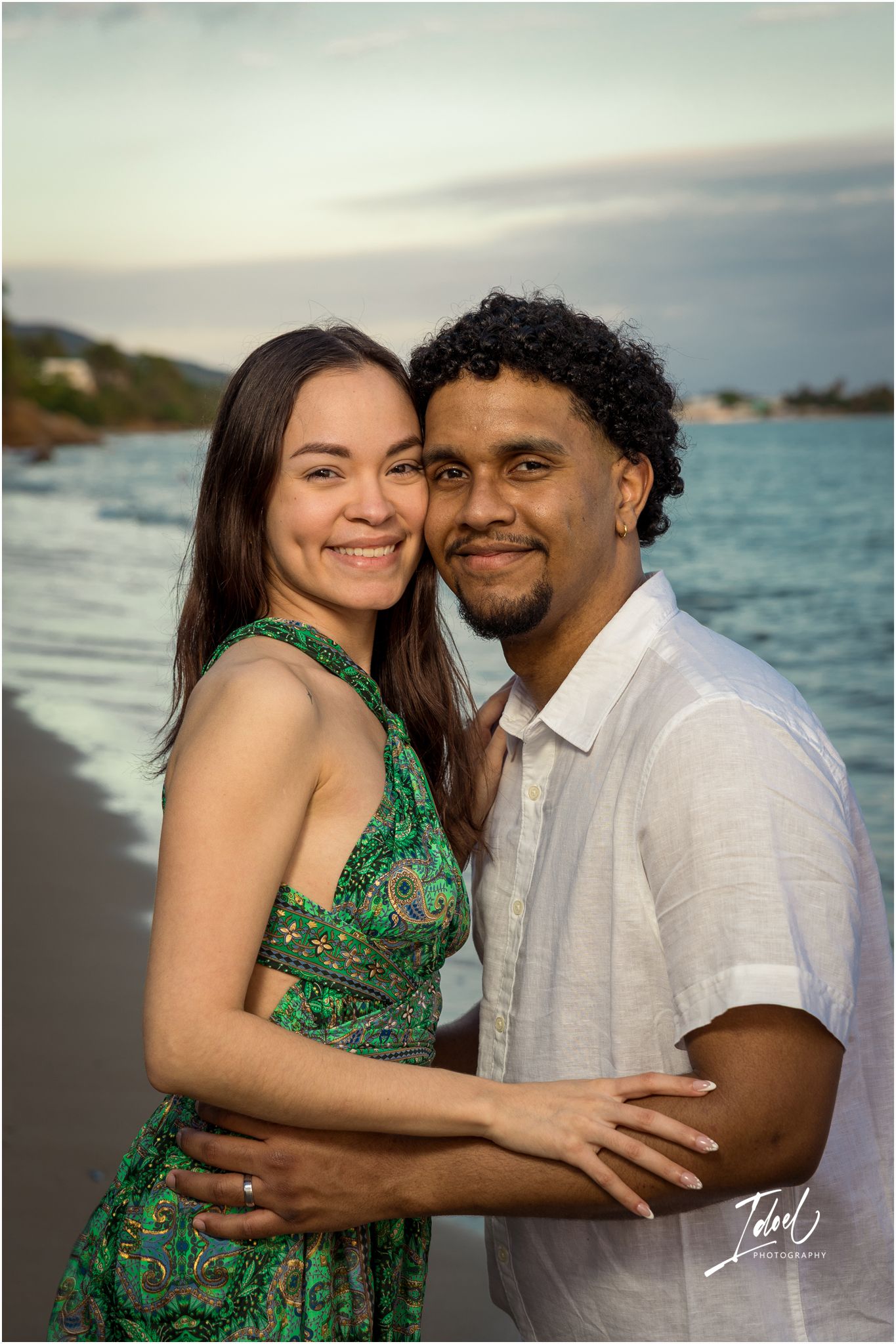 A romantic couple embraces on a beach at sunset, with ocean waves and coastal scenery in the background.
