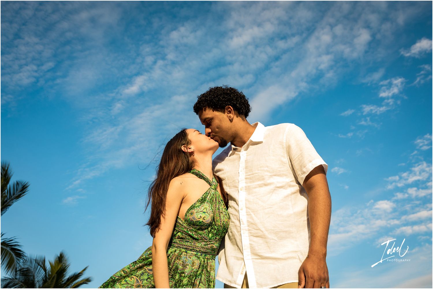A romantic couple shares an intimate moment against a vibrant blue sky with palm trees in the background.