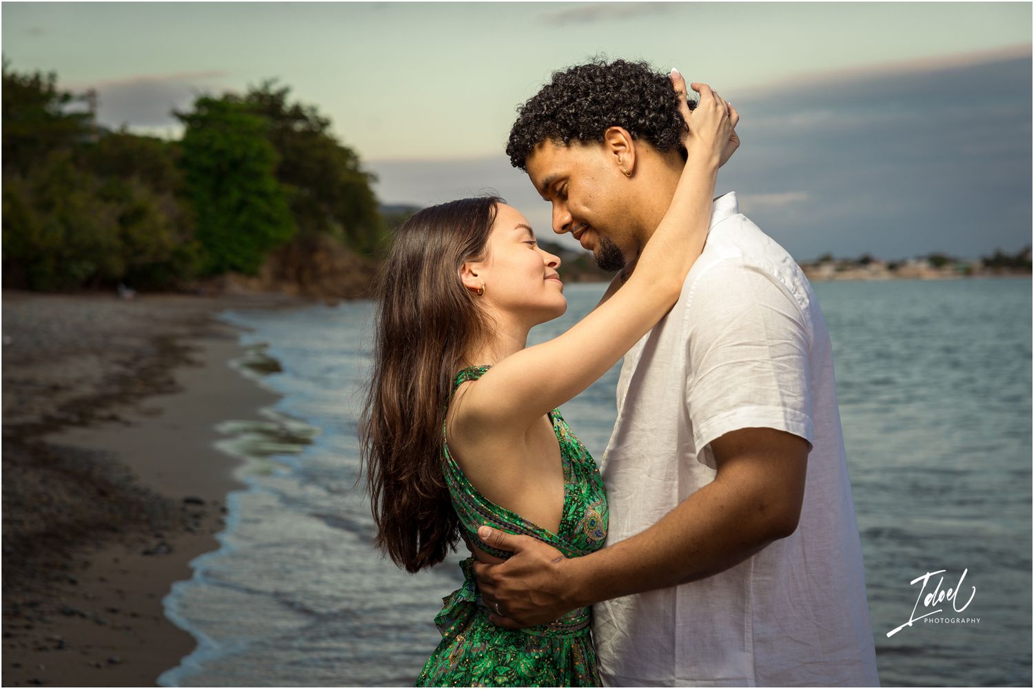 A couple shares an intimate embrace during a beach photoshoot against a coastal backdrop at dusk.