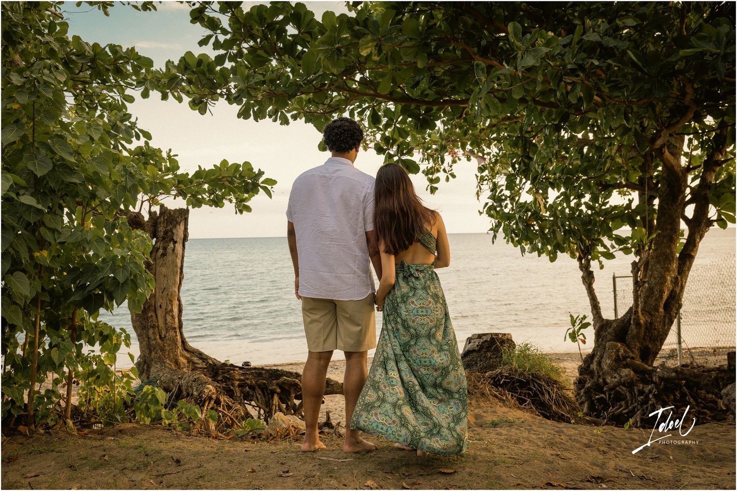 A couple in beach attire stands at the edge of the shore under tropical trees during sunset.