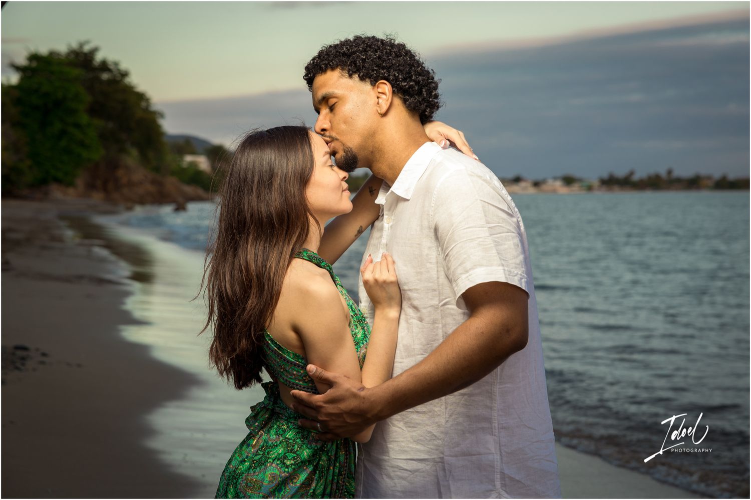 A romantic couple embraces on a beach at sunset with ocean waves and coastline in the background.
