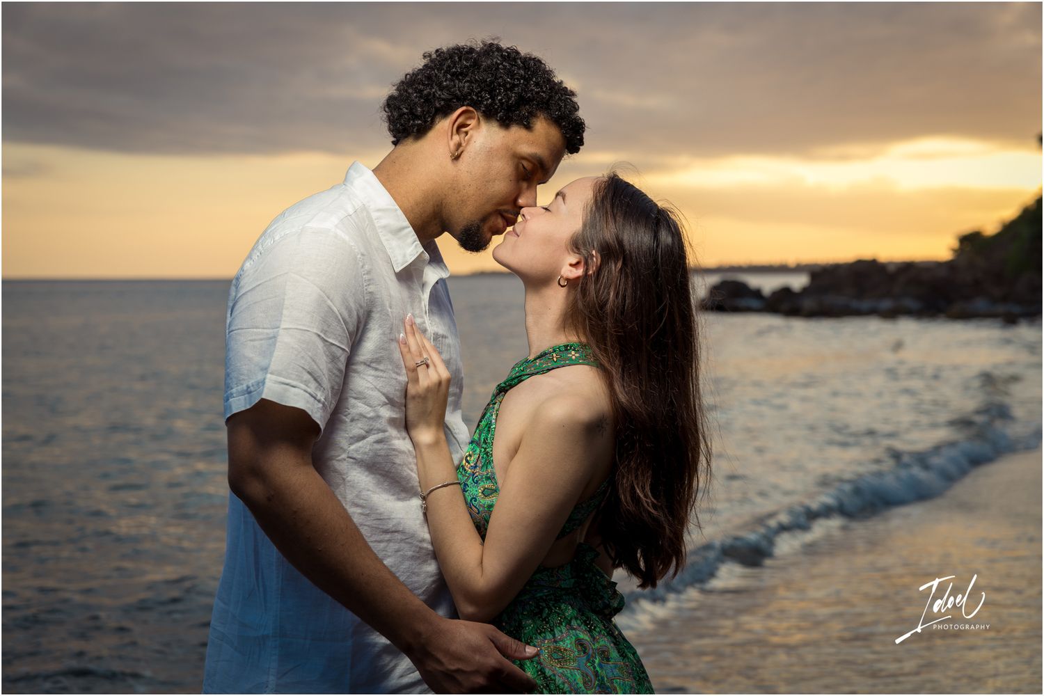 A romantic couple embraces at sunset on a beach shoreline with golden lighting and ocean waves in the background.