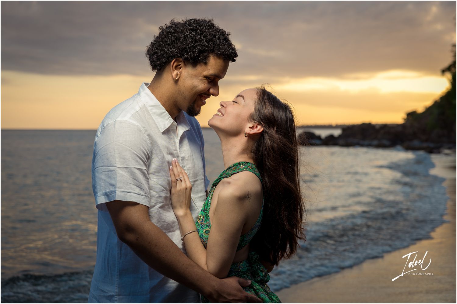Romantic couple embraces on beach at sunset as waves crash against rocky shore in background.