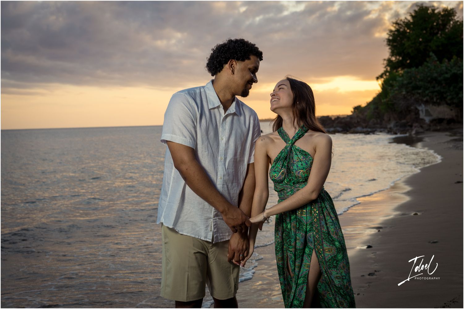 A couple holds hands on a scenic beach during golden sunset with ocean waves and rocky cliffs in the background.