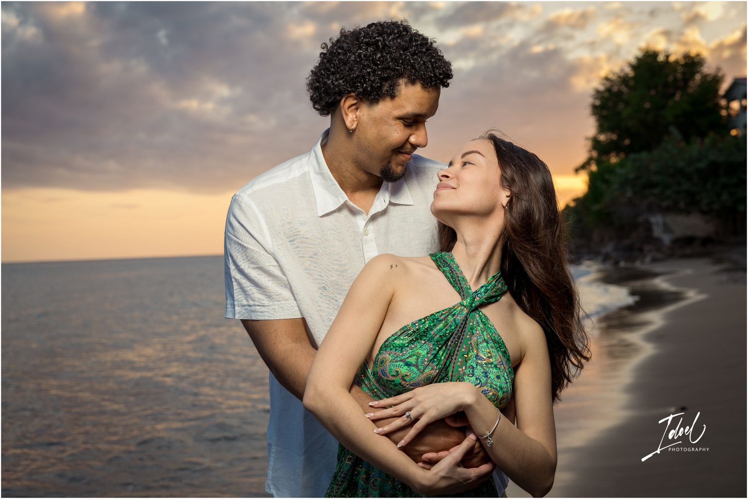 Romantic couple embraces during golden sunset on beach with waves and rocky coastline in background.