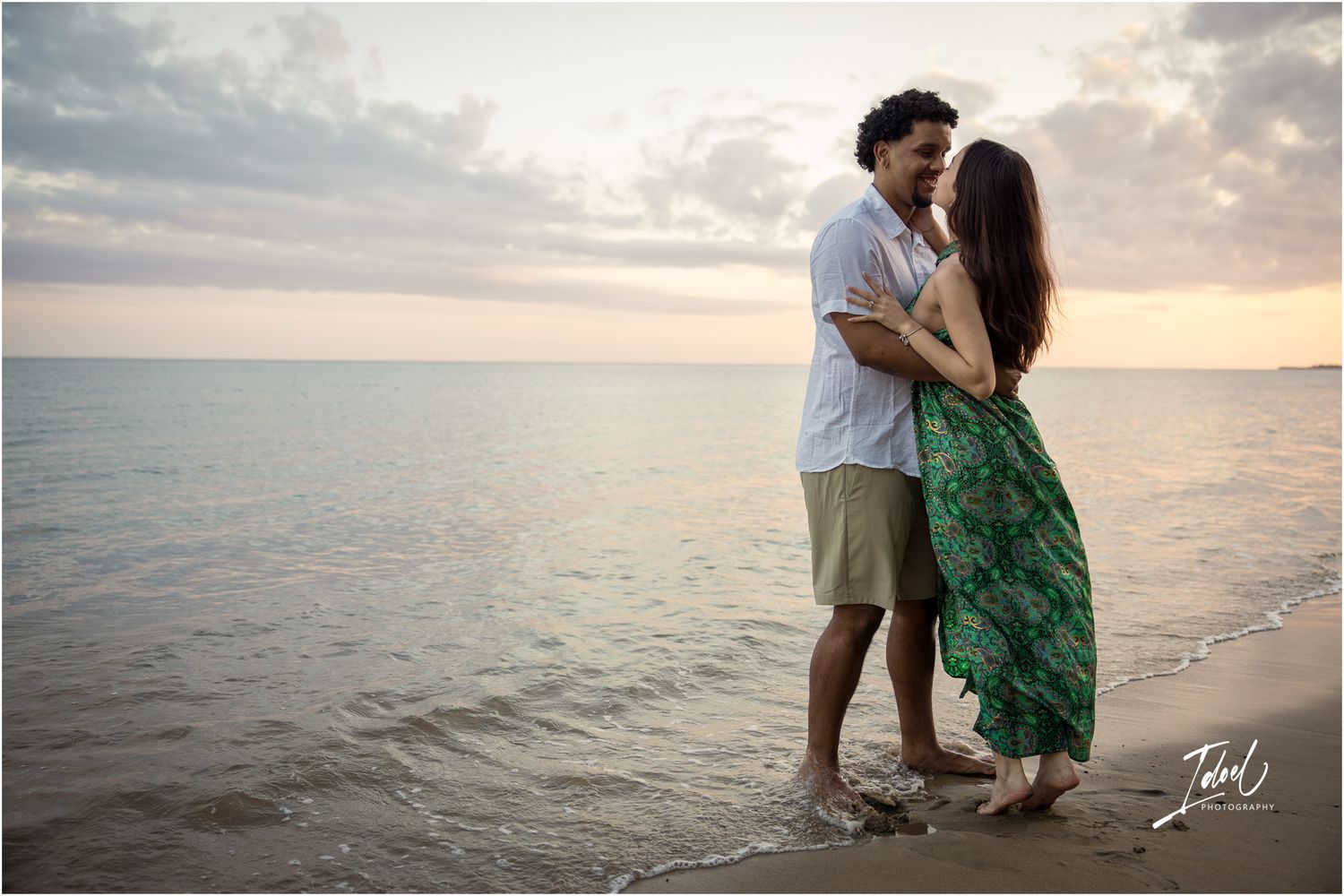 A romantic couple embraces on a beach at sunset with the ocean and cloudy sky in the background.