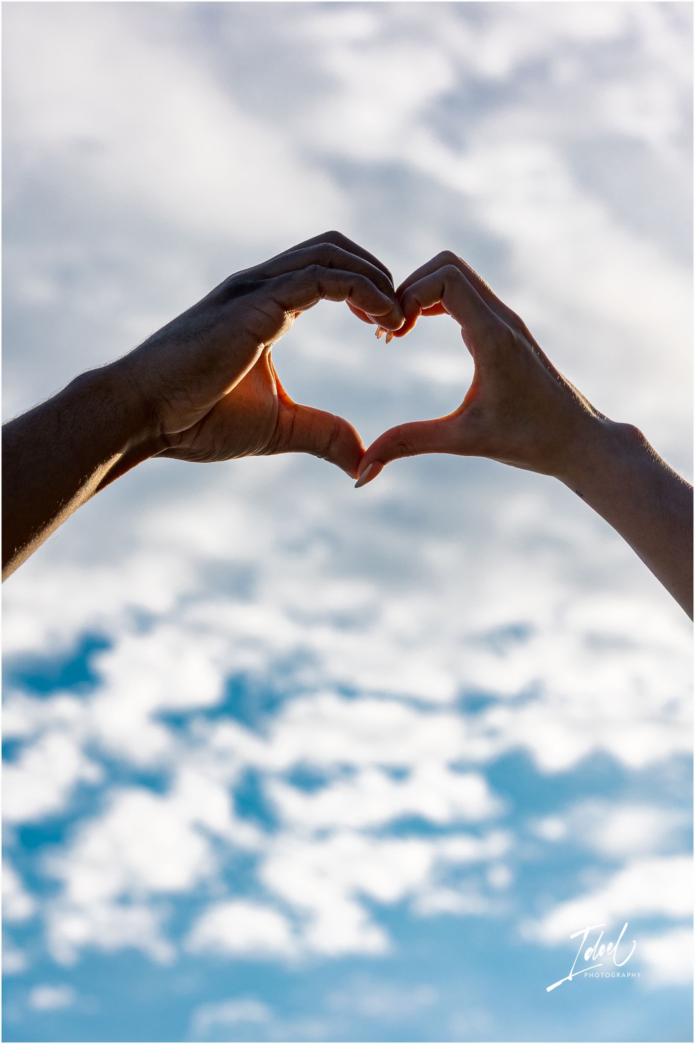 Hands form a heart shape silhouetted against a cloudy blue sky with sunlight peeking through.