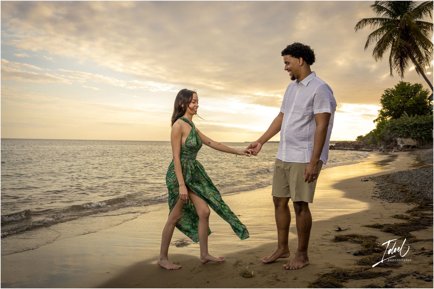 Two people hold hands walking along a tropical beach at sunset with palm trees and ocean in the background.