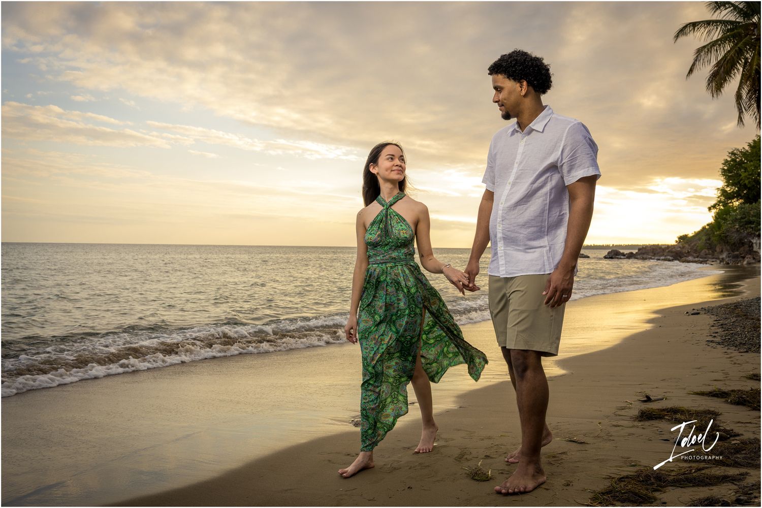 A couple walking along a tropical beach at sunset with palm trees and waves in the background.