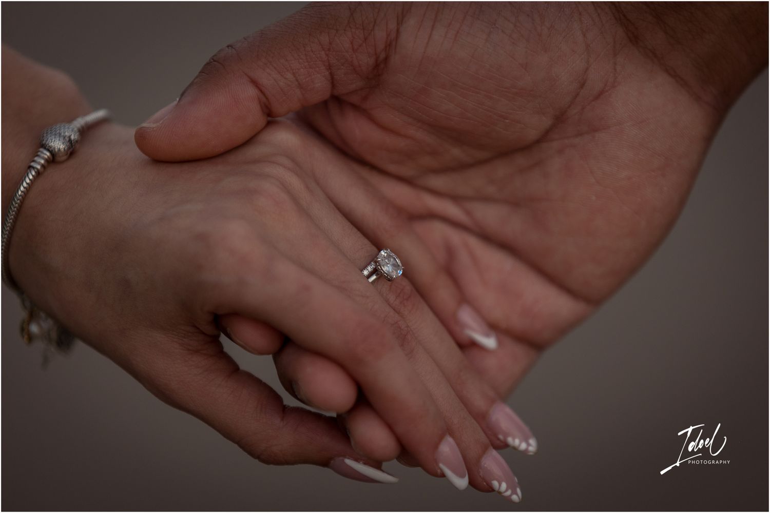 A hand displays an elegant engagement ring with a sparkling center stone against a soft gray background.