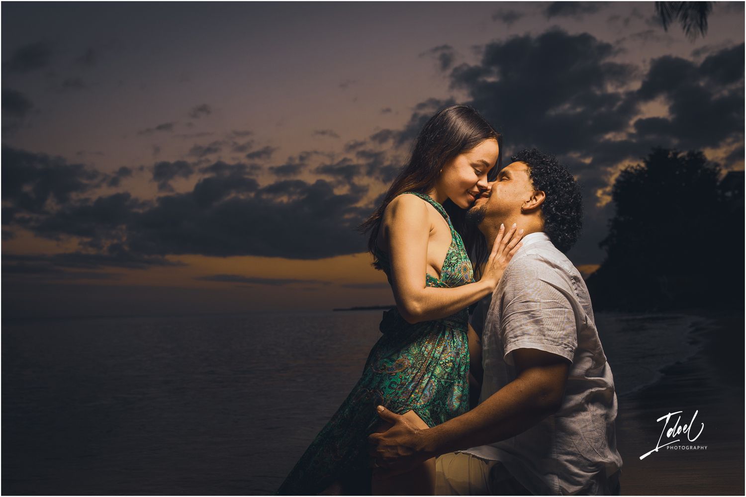 Dramatic sunset silhouettes couple in intimate pose against dark cloudy sky.